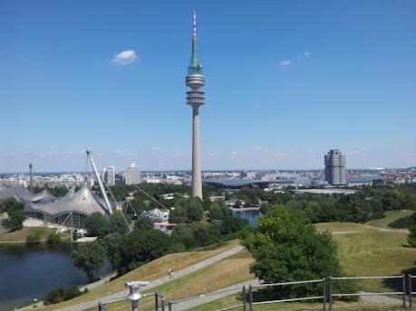 A tall telecommunications tower rises prominently against a clear blue sky, surrounded by modern architectural structures and lush green landscape. A body of water and patches of trees enhance the scenic view, while a distant skyline adds an urban backdrop.