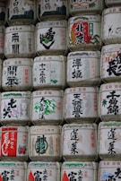 Rows of traditional Japanese sake barrels, intricately decorated with kanji characters and various illustrations. The barrels are made of straw and have a uniform shape, showcasing a blend of cultural and artistic elements.