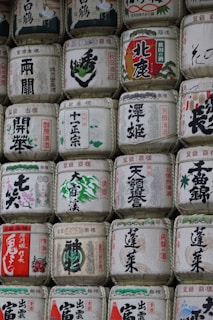Rows of traditional Japanese sake barrels, intricately decorated with kanji characters and various illustrations. The barrels are made of straw and have a uniform shape, showcasing a blend of cultural and artistic elements.