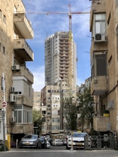 Tall building under construction with a crane on top, surrounded by older residential buildings with air conditioning units. Parked cars line the narrow street, while trees provide some greenery amidst the urban setting. The sky is partly cloudy, with a mix of blue and white.