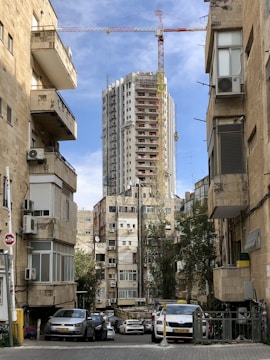 Tall building under construction with a crane on top, surrounded by older residential buildings with air conditioning units. Parked cars line the narrow street, while trees provide some greenery amidst the urban setting. The sky is partly cloudy, with a mix of blue and white.