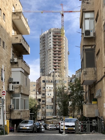 Tall building under construction with a crane on top, surrounded by older residential buildings with air conditioning units. Parked cars line the narrow street, while trees provide some greenery amidst the urban setting. The sky is partly cloudy, with a mix of blue and white.