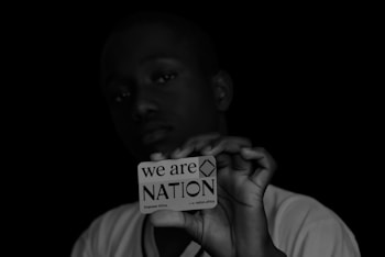 A person holds a card close to the camera, with a focused, serious expression. The card reads 'we are NATION Empower Africa' with a dark background that contrasts with the white text and card. The lighting highlights the card more than the person's face.