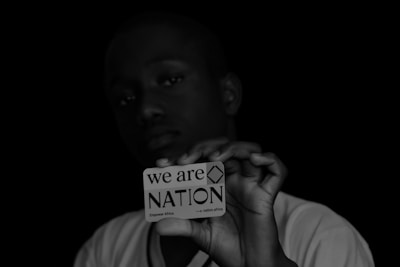 A person holds a card close to the camera, with a focused, serious expression. The card reads 'we are NATION Empower Africa' with a dark background that contrasts with the white text and card. The lighting highlights the card more than the person's face.