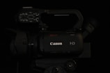 A professional-grade video camera displaying the brand logo on its body. The camera features multiple control knobs and buttons on the top panel, suggesting advanced functionality. The lens and viewfinder are positioned on opposite sides of the camera body, and a handle is attached for ease of movement.