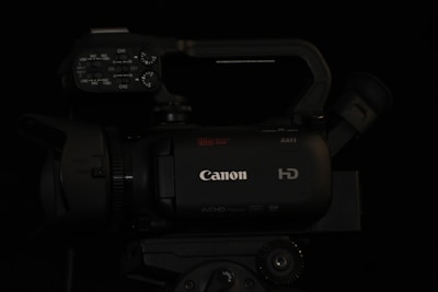 A professional-grade video camera displaying the brand logo on its body. The camera features multiple control knobs and buttons on the top panel, suggesting advanced functionality. The lens and viewfinder are positioned on opposite sides of the camera body, and a handle is attached for ease of movement.