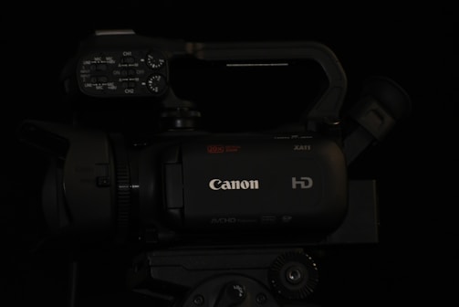 A professional-grade video camera displaying the brand logo on its body. The camera features multiple control knobs and buttons on the top panel, suggesting advanced functionality. The lens and viewfinder are positioned on opposite sides of the camera body, and a handle is attached for ease of movement.