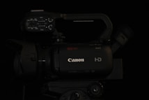 A professional-grade video camera displaying the brand logo on its body. The camera features multiple control knobs and buttons on the top panel, suggesting advanced functionality. The lens and viewfinder are positioned on opposite sides of the camera body, and a handle is attached for ease of movement.
