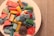 A white plate filled with assorted colorful candies, including various shapes and sizes. The candies are covered in sugar and feature bright colors such as red, yellow, blue, and green. A coiled piece of black candy is also visible.