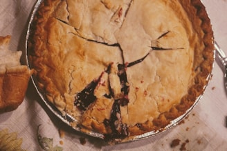 A freshly baked pie with a golden-brown, flaky crust sits on a table, slightly opened to reveal a dark berry filling. A piece of bread rests nearby, and a spoon lies on the tablecloth, suggesting a homemade, rustic setting.