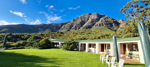 A scenic landscape featuring a lush green lawn in the foreground, bordered by a row of white one-story buildings with a green roof. Beyond the buildings, a verdant hillside and a rugged mountain create a stunning backdrop under a clear blue sky. Several white outdoor chairs and umbrellas are set up on the lawn.