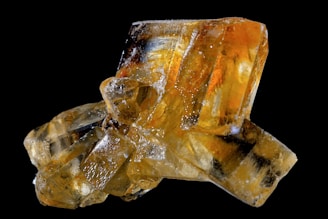 A close-up of a cluster of orange and yellow crystalline structures with a glossy, translucent appearance, set against a black background. The crystals have sharp, angular edges and some display an internal refraction of light, creating a mesmerizing visual effect.