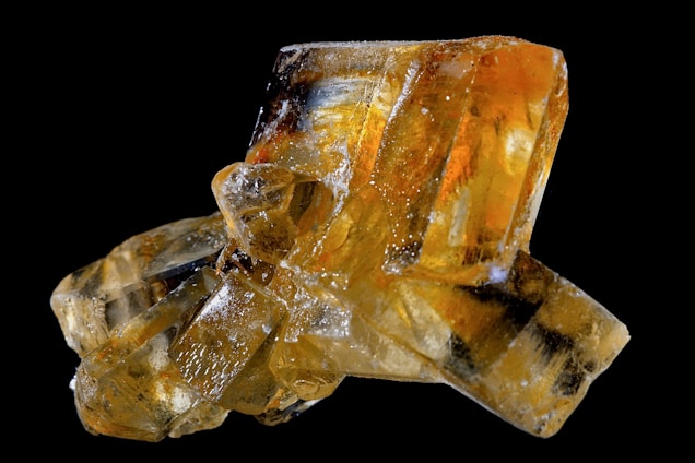 A close-up of a cluster of orange and yellow crystalline structures with a glossy, translucent appearance, set against a black background. The crystals have sharp, angular edges and some display an internal refraction of light, creating a mesmerizing visual effect.