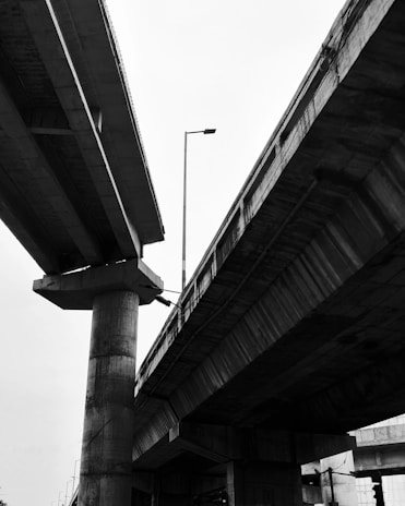 Concrete overpasses intersect against a bright sky. The structures are massive, with visible details of beams and pillars supporting the elevated roads. A lone streetlight stands between the overpasses.