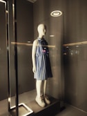 A mannequin is displayed in a glass case illuminated from above by a circular light. It is dressed in a blue and white striped sleeveless dress and white sneakers. The surrounding area has a minimalist and modern design with neutral tones.