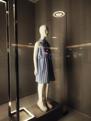 A mannequin is displayed in a glass case illuminated from above by a circular light. It is dressed in a blue and white striped sleeveless dress and white sneakers. The surrounding area has a minimalist and modern design with neutral tones.