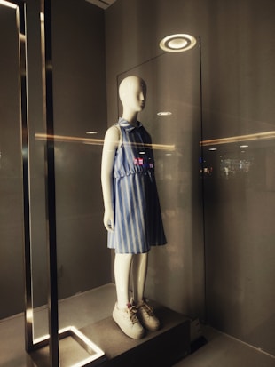 A mannequin is displayed in a glass case illuminated from above by a circular light. It is dressed in a blue and white striped sleeveless dress and white sneakers. The surrounding area has a minimalist and modern design with neutral tones.