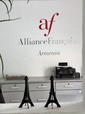 Wall signage featuring the Alliance Française logo in red and grey with the word 'Armenia' below. A minimalist office setting with grey cabinets, a printer on one side, and decorative models of the Eiffel Tower on a desk.