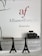 Wall signage featuring the Alliance Française logo in red and grey with the word 'Armenia' below. A minimalist office setting with grey cabinets, a printer on one side, and decorative models of the Eiffel Tower on a desk.