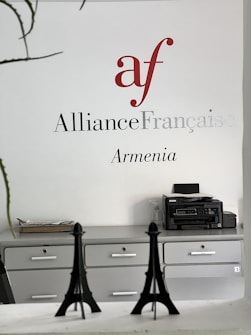 Wall signage featuring the Alliance Française logo in red and grey with the word 'Armenia' below. A minimalist office setting with grey cabinets, a printer on one side, and decorative models of the Eiffel Tower on a desk.