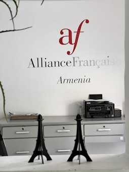 Wall signage featuring the Alliance Française logo in red and grey with the word 'Armenia' below. A minimalist office setting with grey cabinets, a printer on one side, and decorative models of the Eiffel Tower on a desk.
