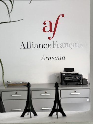 Wall signage featuring the Alliance Française logo in red and grey with the word 'Armenia' below. A minimalist office setting with grey cabinets, a printer on one side, and decorative models of the Eiffel Tower on a desk.