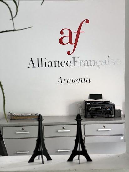Wall signage featuring the Alliance Fran&ccedil;aise logo in red and grey with the word 'Armenia' below. A minimalist office setting with grey cabinets, a printer on one side, and decorative models of the Eiffel Tower on a desk.
