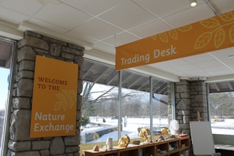 An indoor space with large windows revealing a snowy outdoor scene. The room features stone walls and ceilings with visible panels. Signs with the text 'WELCOME TO THE Nature Exchange' and 'Trading Desk' are prominently displayed. Below the signs are shelves with various items like rocks and shells. The overall setting suggests a nature or educational theme.