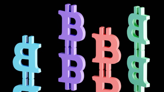 Multiple Bitcoin symbols in different colors are arranged vertically against a black background. These symbols appear in blue, purple, red, and green, with a reflective surface below each one, creating a mirrored effect.