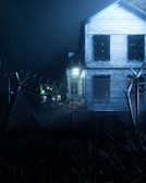 A two-story wooden house with boarded-up windows stands behind a barbed-wire fence. The atmosphere is dark and eerie, with dim lighting illuminating a green jeep parked beside the house. The scene is surrounded by overgrown grass, adding to the abandoned feel.