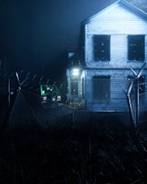 A two-story wooden house with boarded-up windows stands behind a barbed-wire fence. The atmosphere is dark and eerie, with dim lighting illuminating a green jeep parked beside the house. The scene is surrounded by overgrown grass, adding to the abandoned feel.