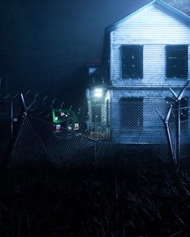 A two-story wooden house with boarded-up windows stands behind a barbed-wire fence. The atmosphere is dark and eerie, with dim lighting illuminating a green jeep parked beside the house. The scene is surrounded by overgrown grass, adding to the abandoned feel.