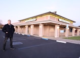 A man stands in an empty parking lot in front of a large, beige-colored building with multiple signs displaying MEALPRO.COM. The building has illuminated lights along its exterior, creating a warm glow. The parking lot features marked parking spaces, a reserved parking sign, and some landscaping.