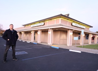 A man stands in an empty parking lot in front of a large, beige-colored building with multiple signs displaying MEALPRO.COM. The building has illuminated lights along its exterior, creating a warm glow. The parking lot features marked parking spaces, a reserved parking sign, and some landscaping.