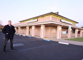 A man stands in an empty parking lot in front of a large, beige-colored building with multiple signs displaying MEALPRO.COM. The building has illuminated lights along its exterior, creating a warm glow. The parking lot features marked parking spaces, a reserved parking sign, and some landscaping.