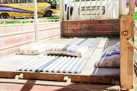 A flatbed truck is loaded with corrugated metal sheets and several bags of construction materials, likely cement or a similar substance. The truck has open sides and a rustic, weathered appearance. In the background, a yellow bus with blue accents is parked, and greenery can be seen.