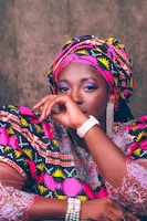 A person is dressed in vibrant traditional attire featuring bold patterns and colors. The outfit includes a matching headwrap and clothing with intricate geometric designs in pink, yellow, black, and white. The individual is wearing accessories such as a pearl bracelet, a stylish watch, and eye-catching dangling earrings. The facial expression conveys a calm and confident presence.