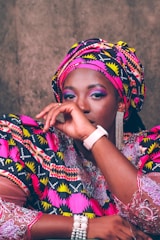 A person is dressed in vibrant traditional attire featuring bold patterns and colors. The outfit includes a matching headwrap and clothing with intricate geometric designs in pink, yellow, black, and white. The individual is wearing accessories such as a pearl bracelet, a stylish watch, and eye-catching dangling earrings. The facial expression conveys a calm and confident presence.
