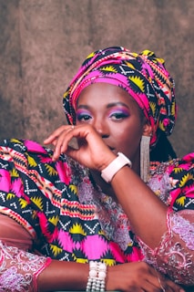 A person is dressed in vibrant traditional attire featuring bold patterns and colors. The outfit includes a matching headwrap and clothing with intricate geometric designs in pink, yellow, black, and white. The individual is wearing accessories such as a pearl bracelet, a stylish watch, and eye-catching dangling earrings. The facial expression conveys a calm and confident presence.