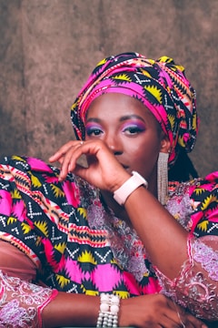 A person is dressed in vibrant traditional attire featuring bold patterns and colors. The outfit includes a matching headwrap and clothing with intricate geometric designs in pink, yellow, black, and white. The individual is wearing accessories such as a pearl bracelet, a stylish watch, and eye-catching dangling earrings. The facial expression conveys a calm and confident presence.