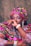 A person is dressed in vibrant traditional attire featuring bold patterns and colors. The outfit includes a matching headwrap and clothing with intricate geometric designs in pink, yellow, black, and white. The individual is wearing accessories such as a pearl bracelet, a stylish watch, and eye-catching dangling earrings. The facial expression conveys a calm and confident presence.