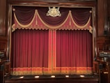 An ornate theater stage with rich burgundy curtains adorned with gold tassels and decorative patterns. The emblem above the curtains adds a touch of elegance to the classical setting. Wooden panels frame the sides, enhancing the traditional atmosphere.