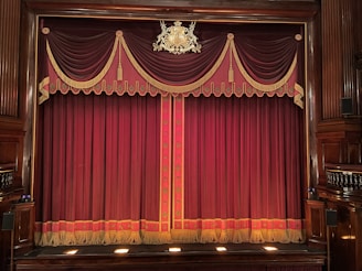 An ornate theater stage with rich burgundy curtains adorned with gold tassels and decorative patterns. The emblem above the curtains adds a touch of elegance to the classical setting. Wooden panels frame the sides, enhancing the traditional atmosphere.