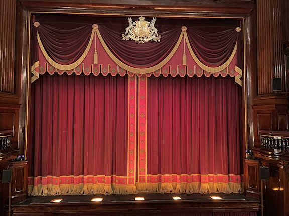 An ornate theater stage with rich burgundy curtains adorned with gold tassels and decorative patterns. The emblem above the curtains adds a touch of elegance to the classical setting. Wooden panels frame the sides, enhancing the traditional atmosphere.