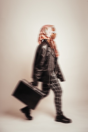 A person wearing a hijab, sunglasses, and a leather jacket strides confidently with a black briefcase. The image has a blurred motion effect, giving a sense of movement.
