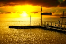 A breathtaking golden sunset over a calm sea, with the sky painted in vibrant hues of yellow and orange. A wooden dock extends into the water, bearing a sign that reads 'Majestic Barri&egrave;re'. A lighthouse stands on a rocky outcrop in the distance.