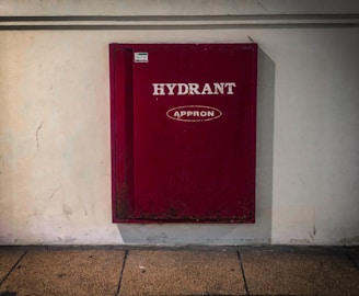 A red fire hydrant cabinet is mounted on a plain white wall. The cabinet is rectangular and labeled with the text 'HYDRANT' and 'APPRON' in white lettering. The floor appears to be a textured, tan-colored concrete.