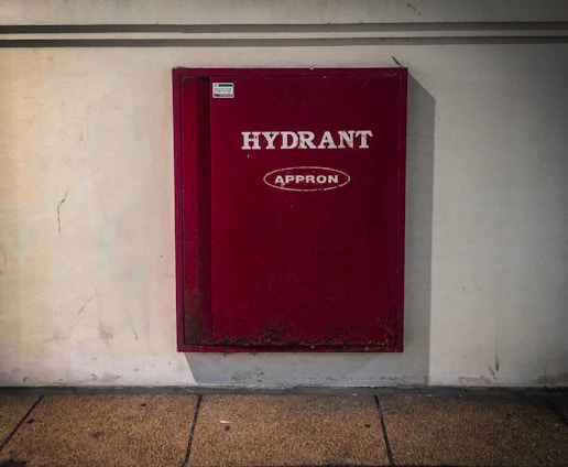 A red fire hydrant cabinet is mounted on a plain white wall. The cabinet is rectangular and labeled with the text 'HYDRANT' and 'APPRON' in white lettering. The floor appears to be a textured, tan-colored concrete.
