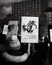 A black-and-white photograph depicting a crowd of people holding protest signs. One prominent sign features an artistic drawing of a woman in a defiant pose, with the words 'Woman, Life, Freedom, Iran' written beneath it. The crowd is gathered outdoors, and a tall building can be seen in the background.