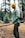 A person stands in a forested area, wearing a yellow beanie, green jacket, and dark pants. They have a colorful backpack and carry an axe attached to their belt. The background features evergreen trees and the ground is covered with light patches of snow and fallen branches.